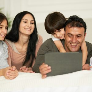 Things to do at home. Happy latin family with cute little kids using digital tablet lying on the bed